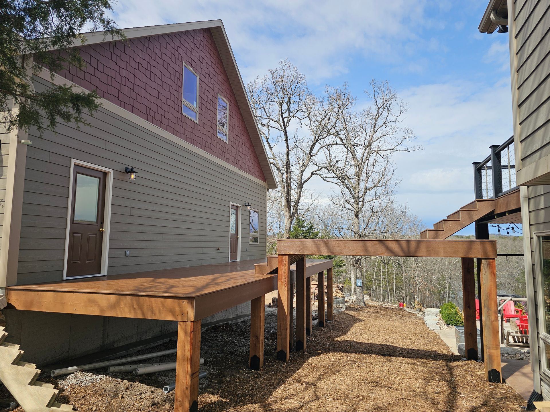 Wooden deck attached to a two-story house with a pathway and pergola in a hillside setting.