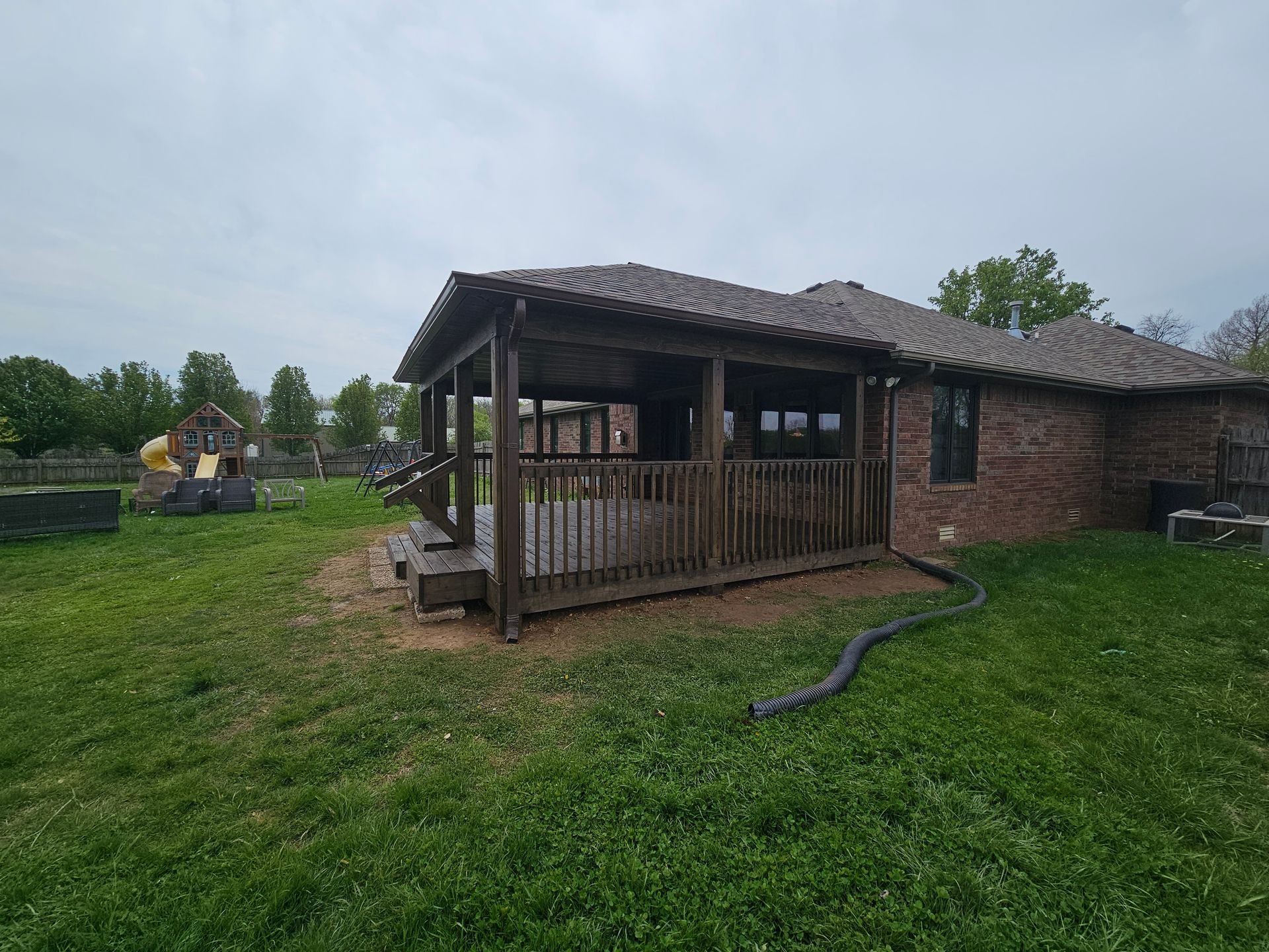 Dark wooden deck and covered patio attached to a brick house on a grassy lawn.