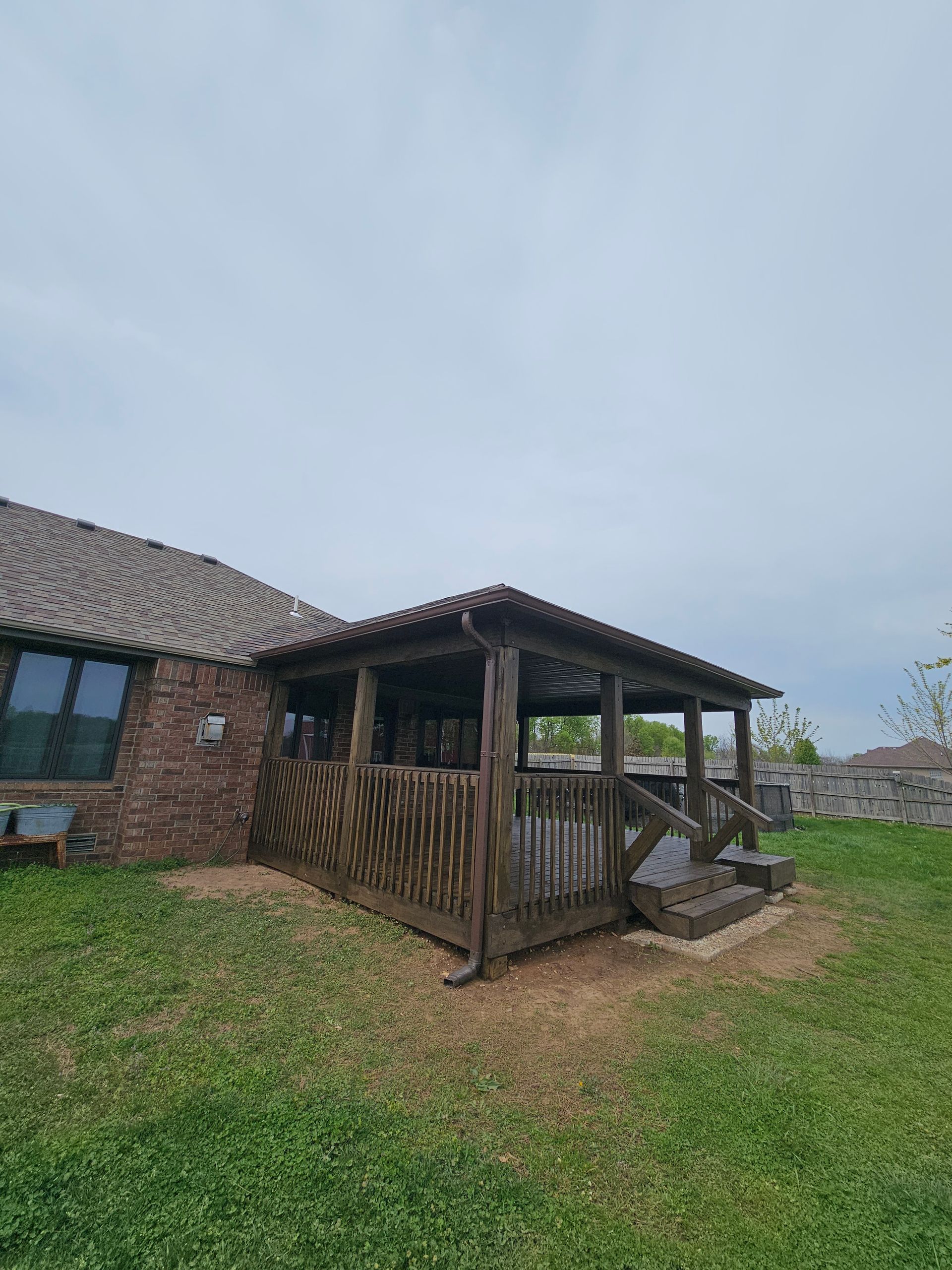 Wooden gazebo with dark tiled roof attached to a brick building, on a grassy lawn under a cloudy sky.