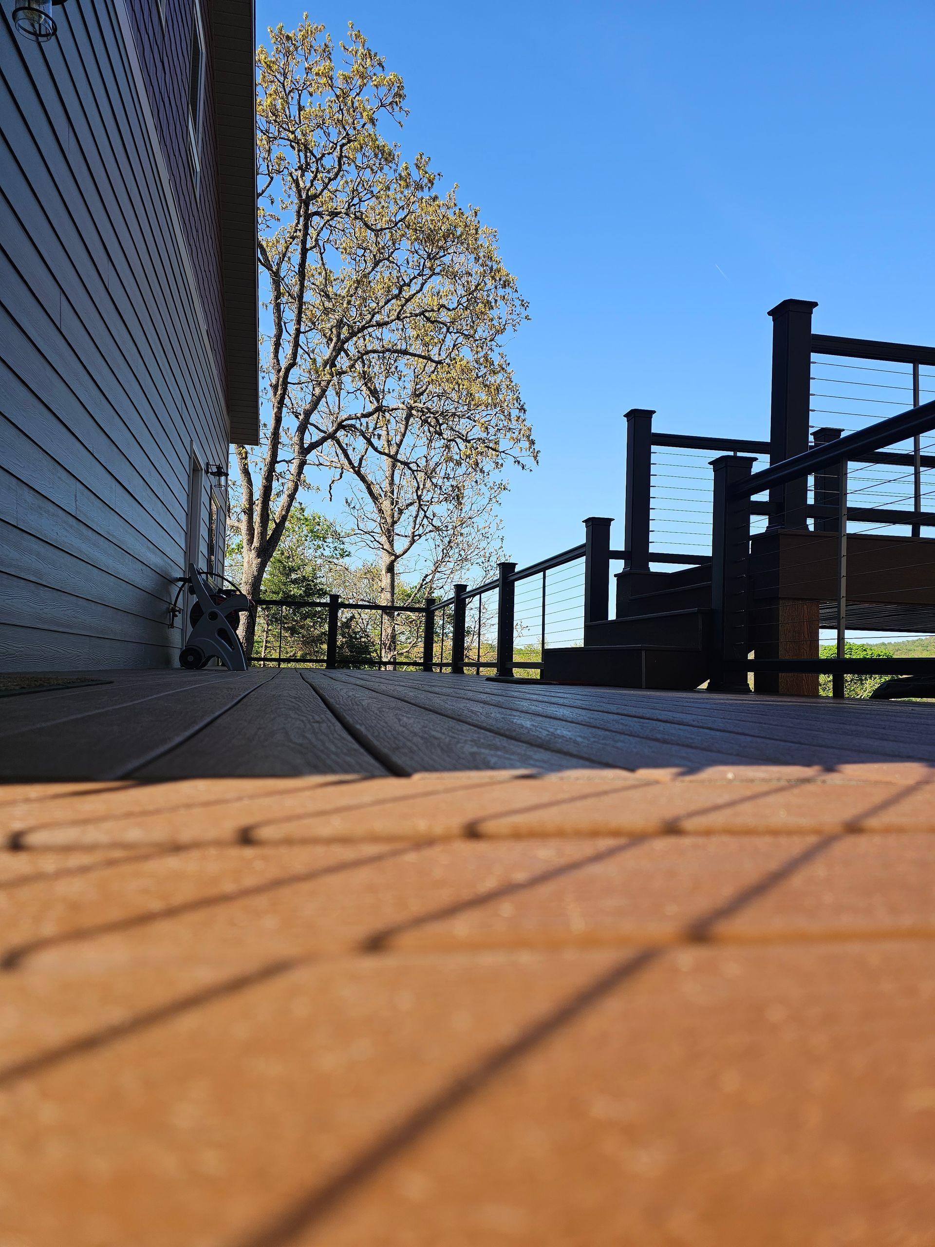 Low-angle view of a wooden deck with dark railing, a building on the left, and a tree against a clear blue sky.