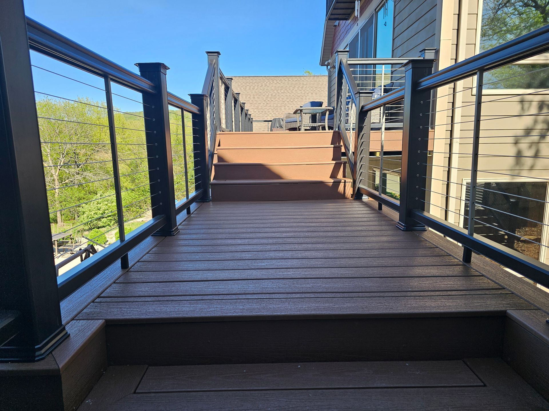Wooden deck and stairs with black railing and cable infill, leading up to a house on a sunny day.