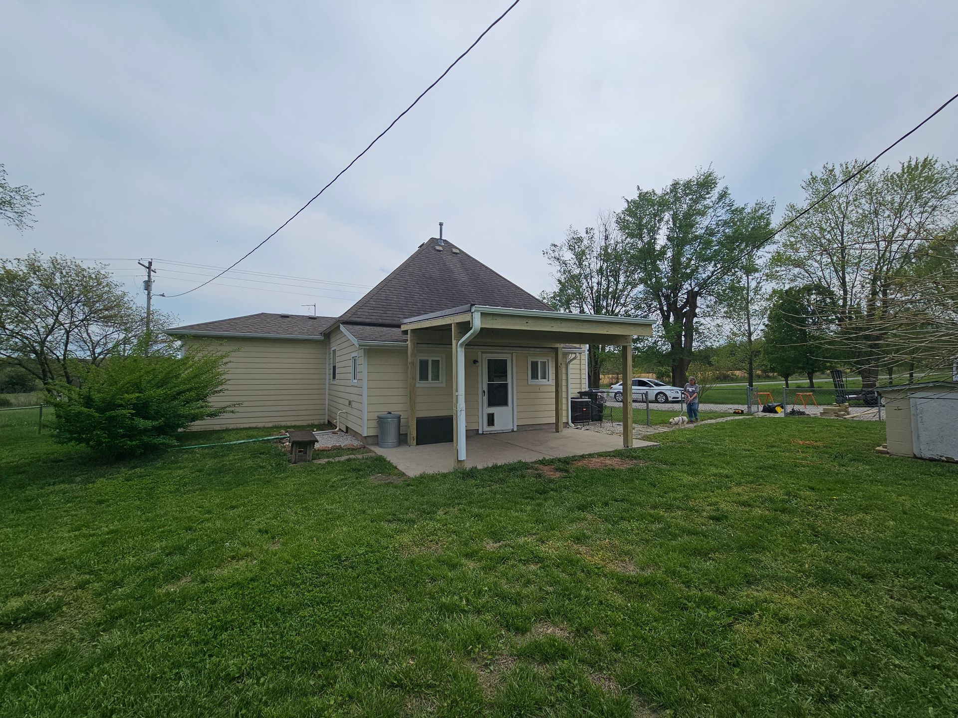 Small beige house with a covered porch and round roof. Green grass and trees under a cloudy sky.