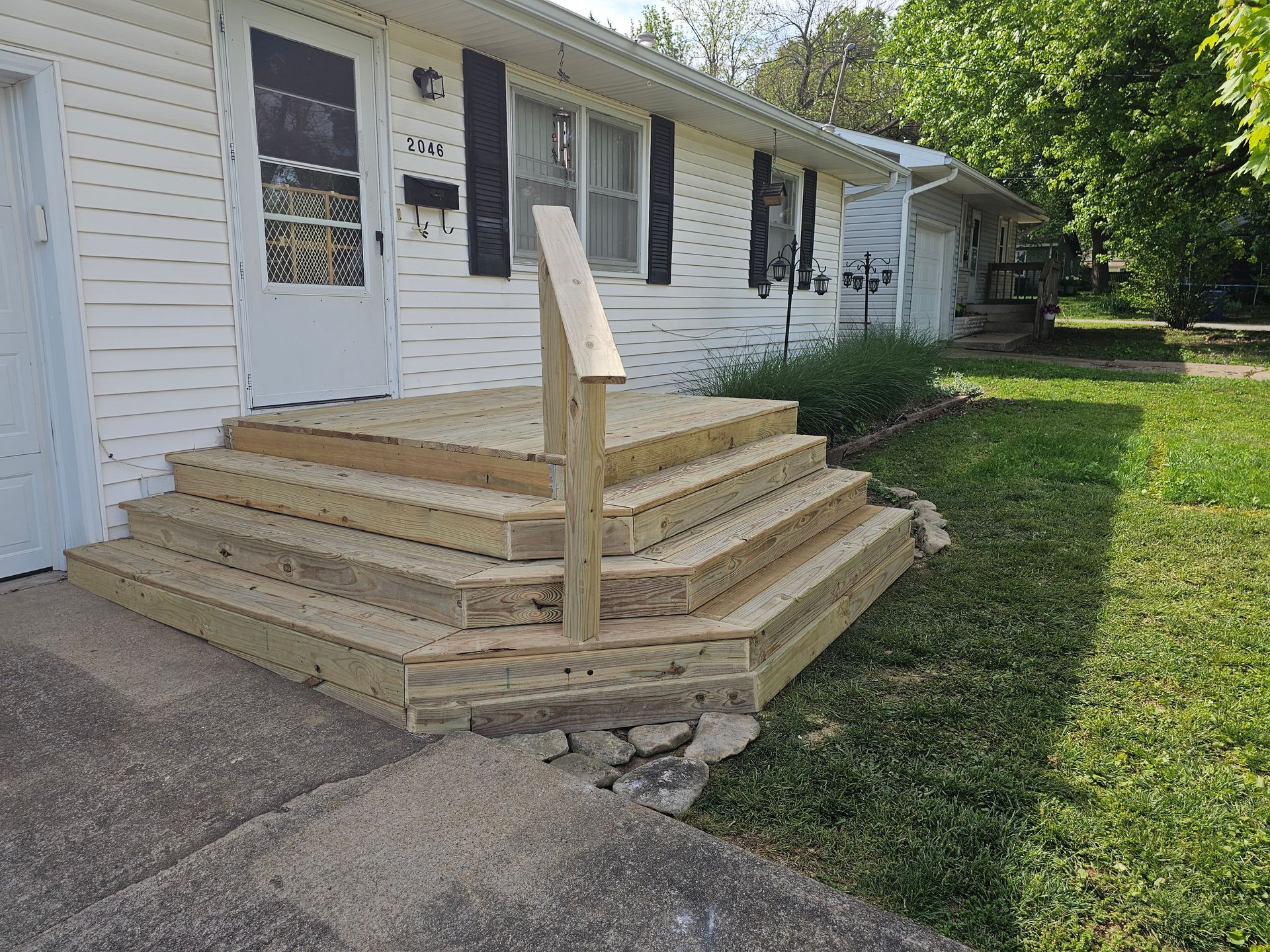 Wooden steps leading up to a white house with a handrail. Green grass surrounds the steps and concrete sidewalk.