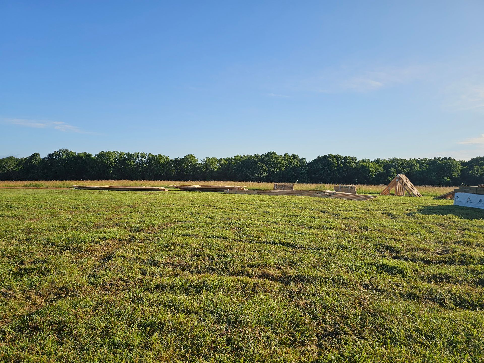 Field with construction materials, trees in the distance under a blue sky.