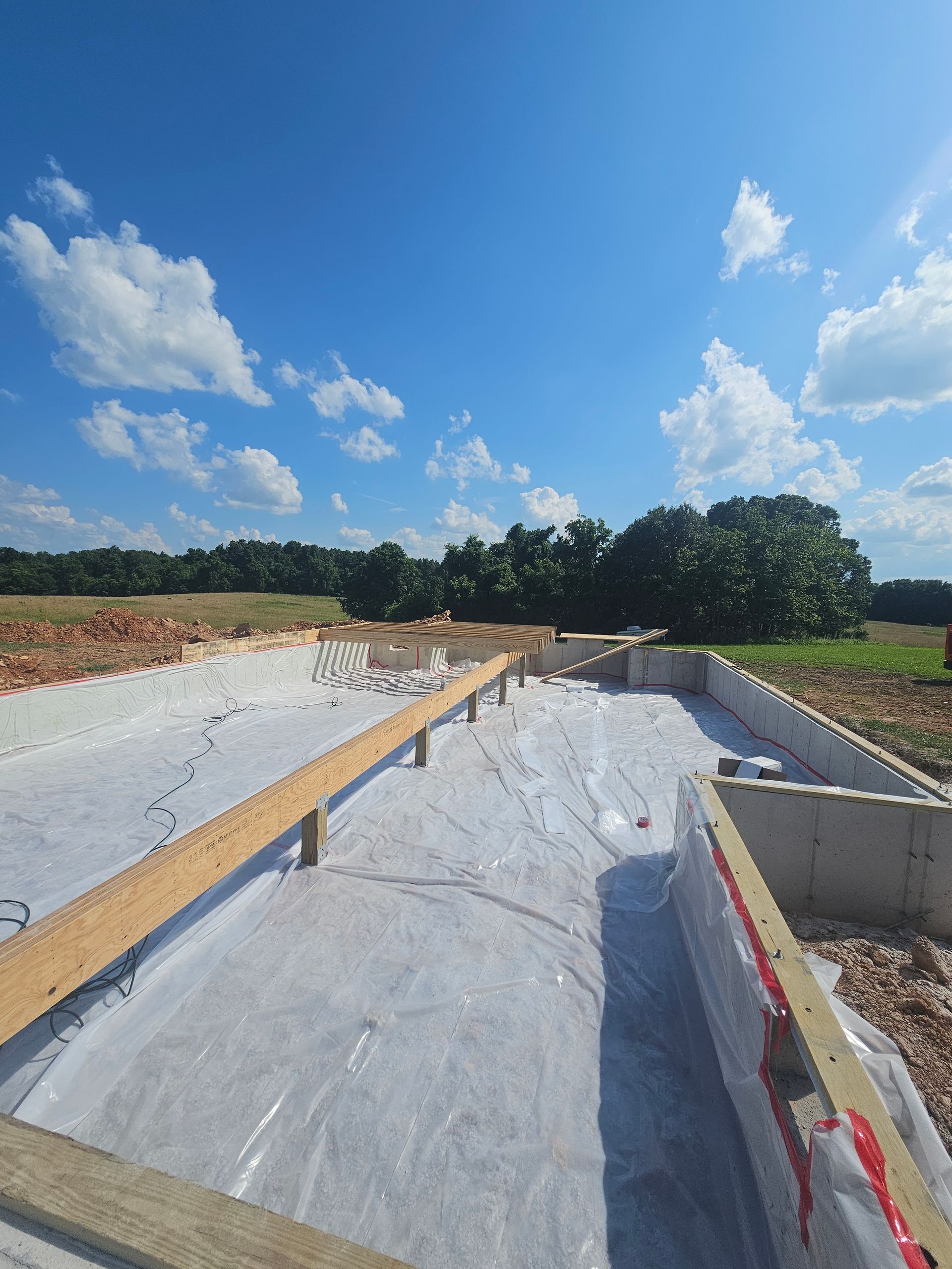 Construction site: concrete foundation with wooden beams and white plastic sheeting, blue sky with clouds.