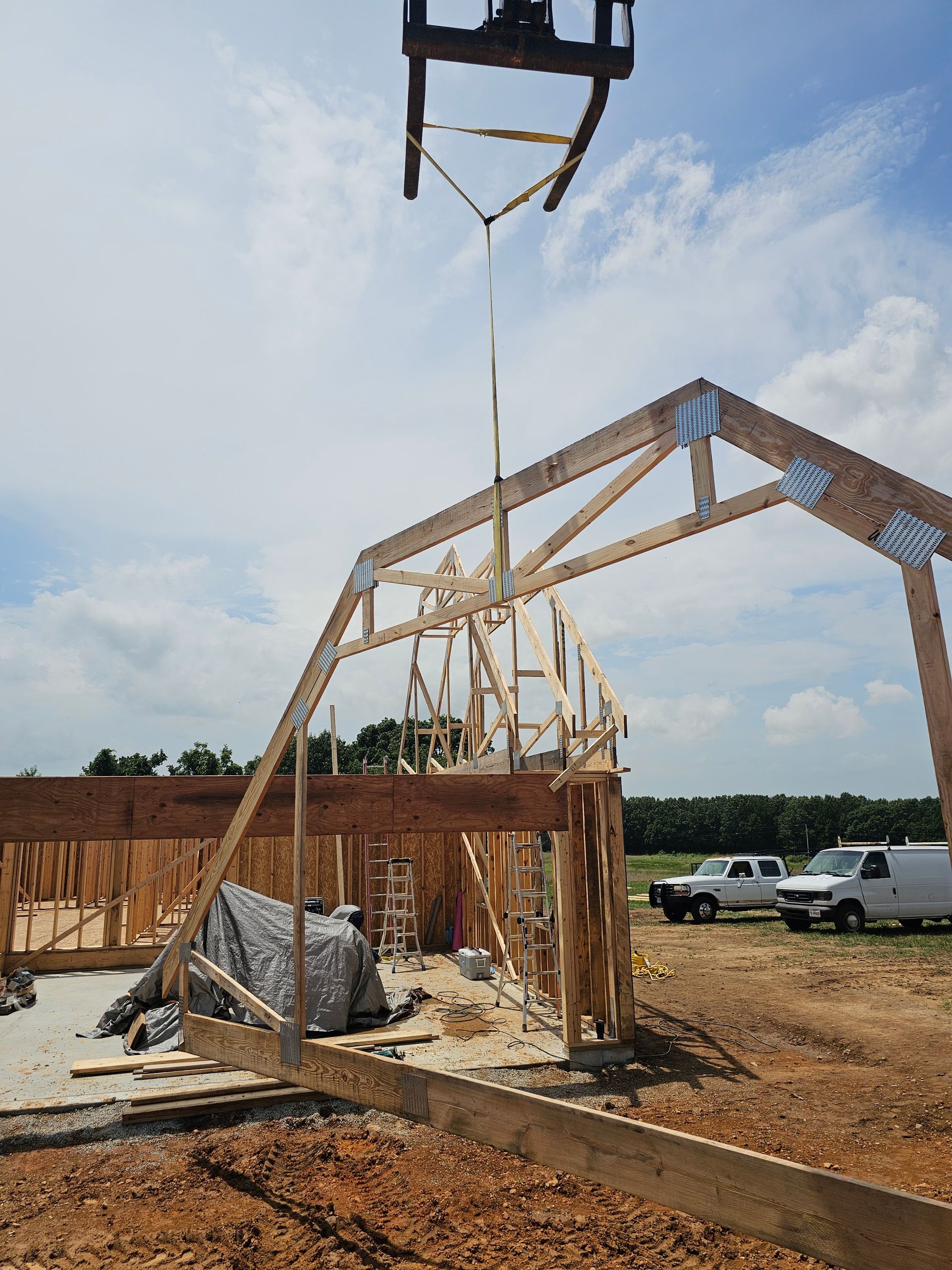 Forklift lifting a wooden roof truss onto a partially constructed building frame on a sunny construction site.