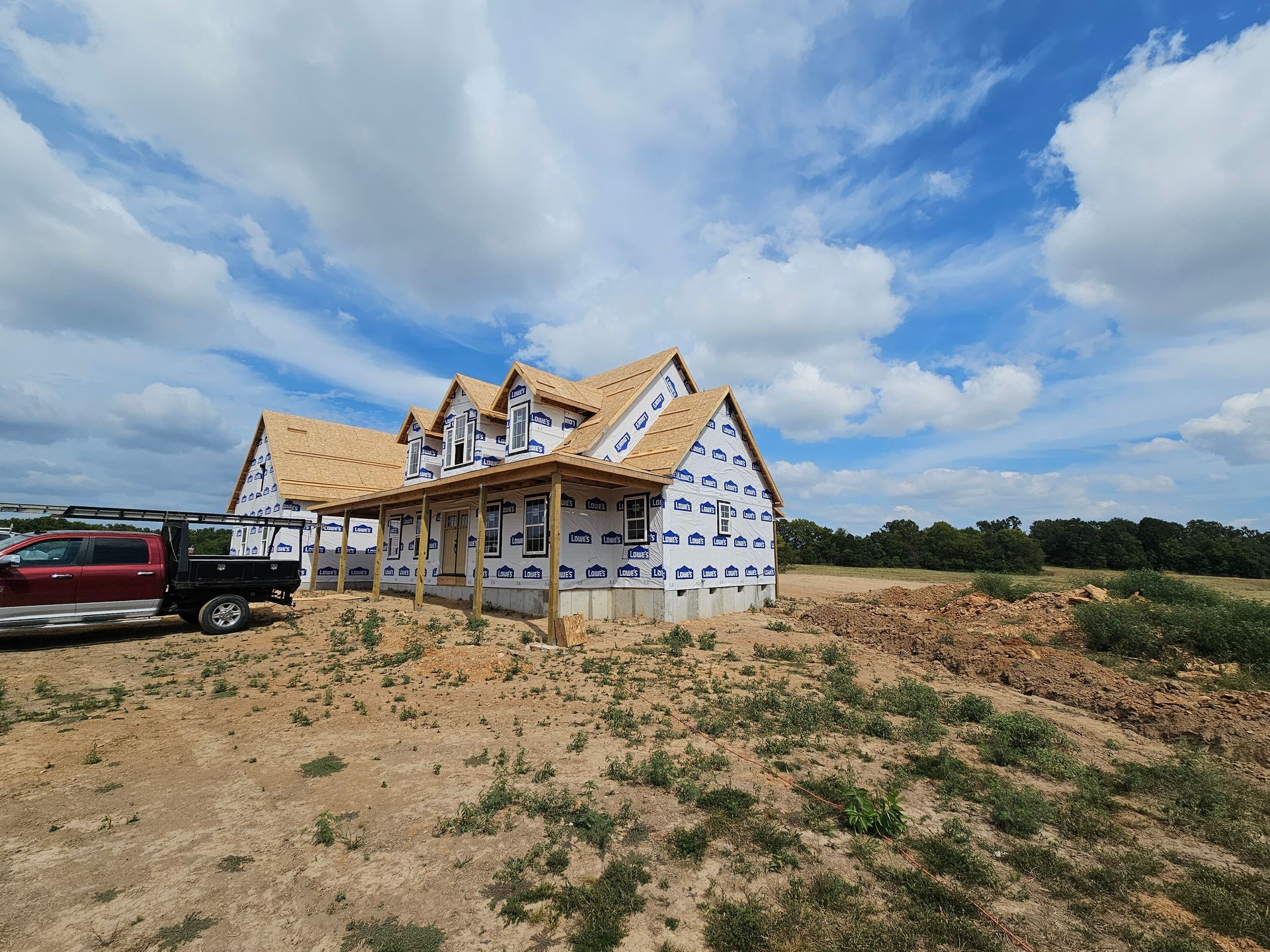 House under construction with blue wrap, brown roof, and red truck parked nearby under a partly cloudy sky.