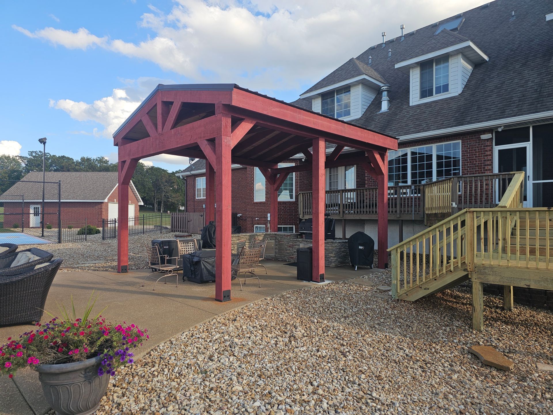 Red wooden pergola next to a house with a deck. The sky is blue and the ground is covered in rocks.