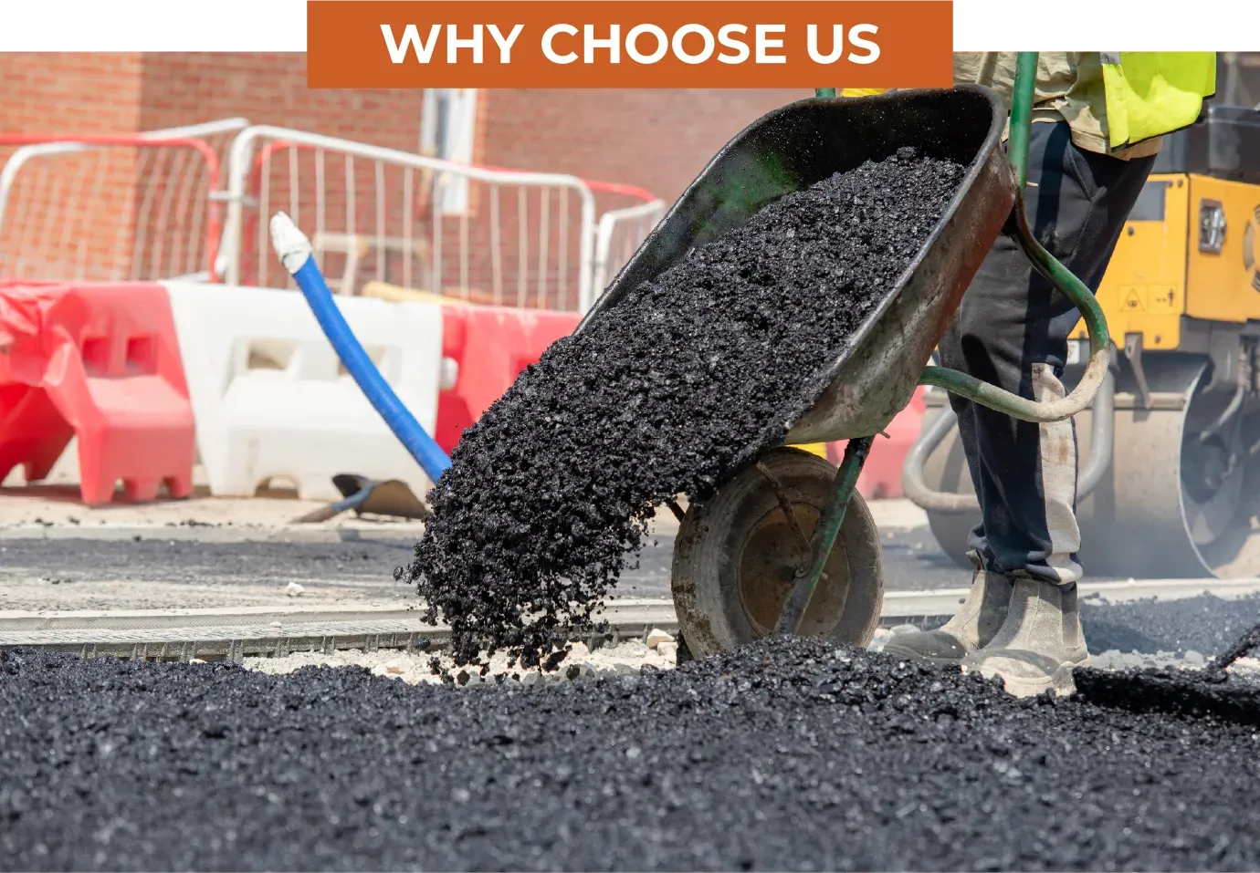 A worker pours hot asphalt from a wheelbarrow onto a road surface at a construction site with heavy machinery nearby.