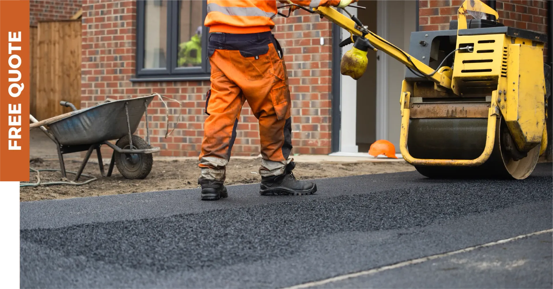 A construction worker uses a yellow steamroller to pave a patch of asphalt in front of a brick building.