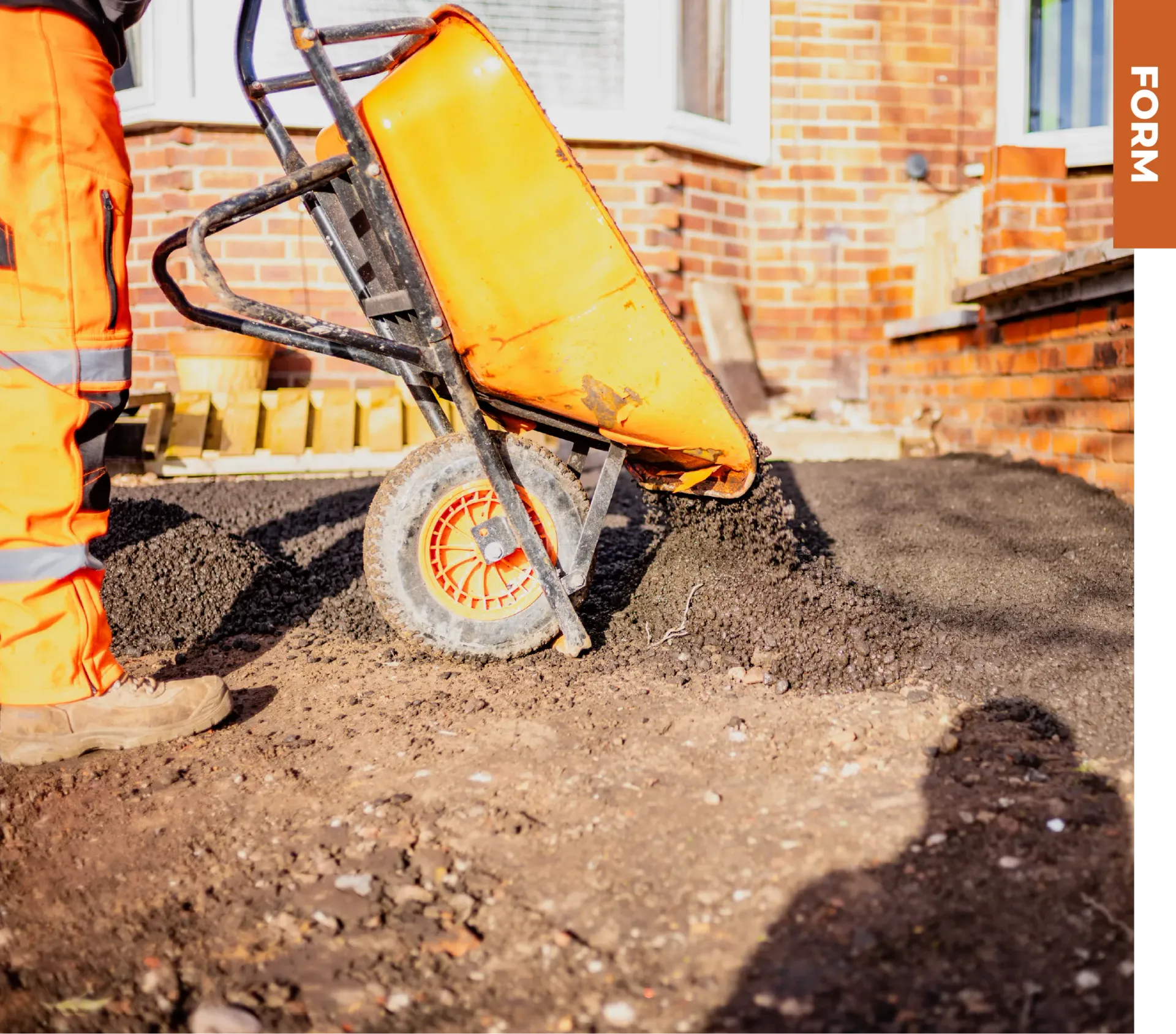 A person in high-visibility orange work trousers dumps gravel from a yellow wheelbarrow onto a dirt construction site.