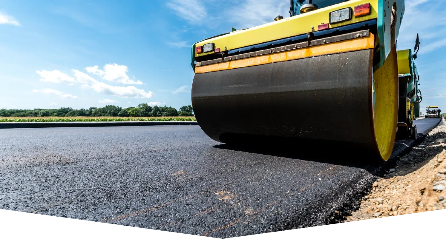 A bright yellow steamroller compresses freshly laid, dark asphalt on a road construction site under a sunny blue sky.