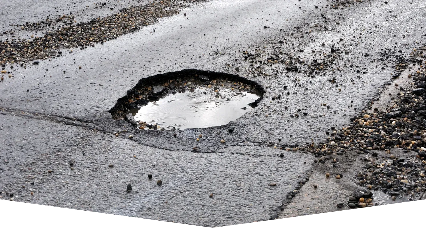 A large pothole filled with stagnant water on a cracked and damaged asphalt road surface.