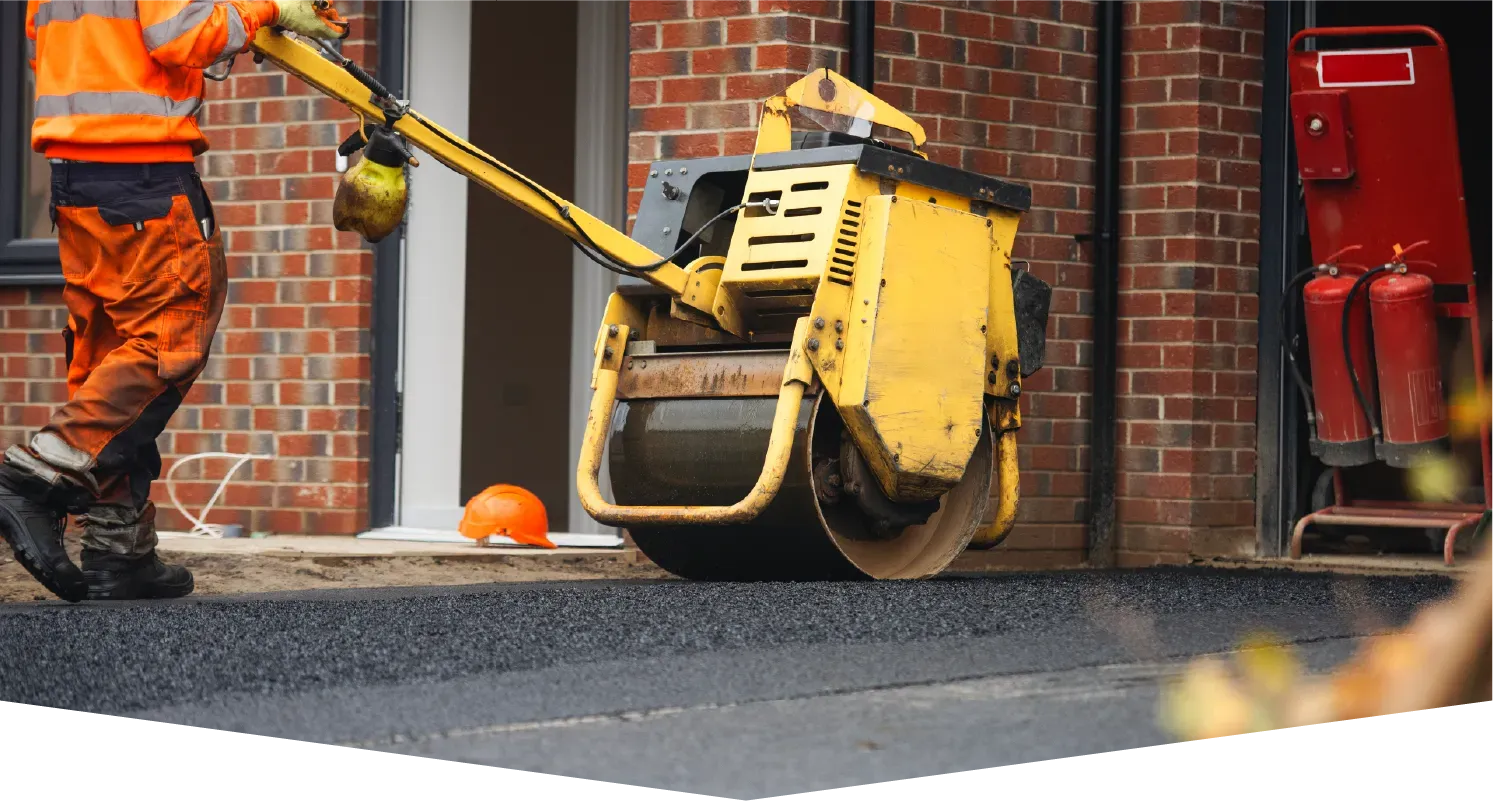 A construction worker in high-visibility gear uses a yellow walk-behind roller to compact fresh asphalt on a driveway.
