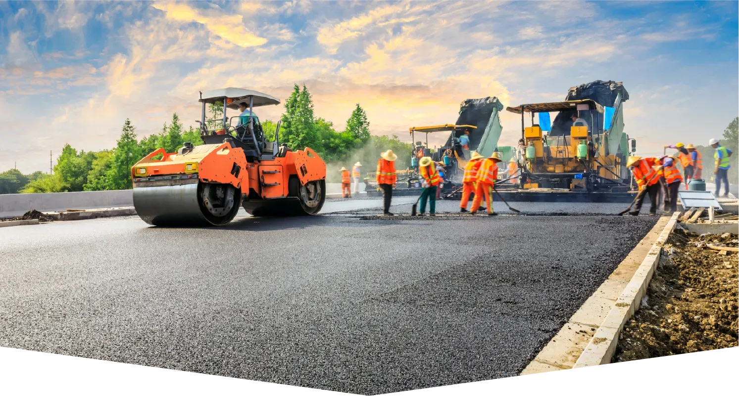 Road construction crew members working with paving machinery on a freshly laid asphalt road under a sunset sky.