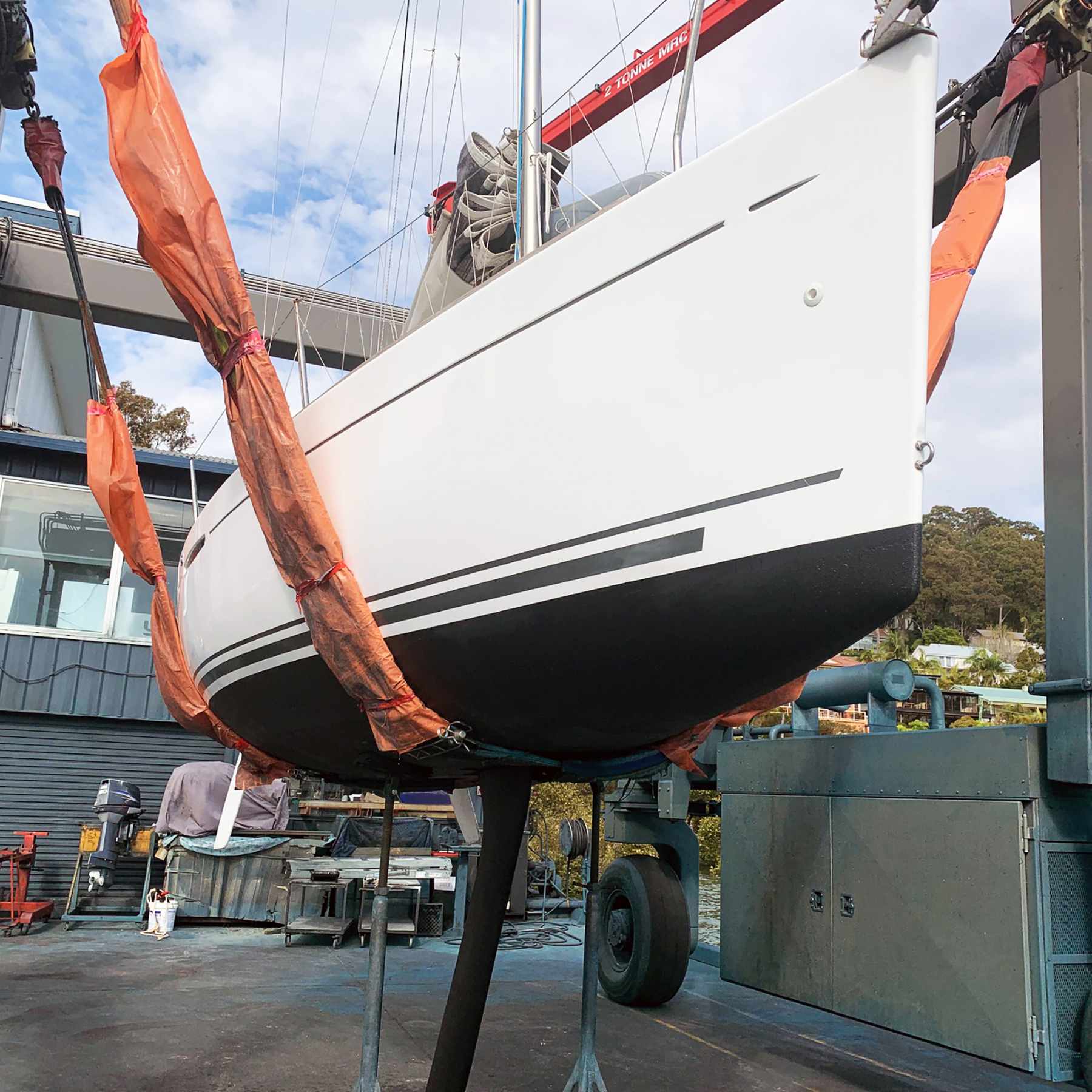 White sailboat being lifted by crane slings. Black hull, grey stripes, blue sky.