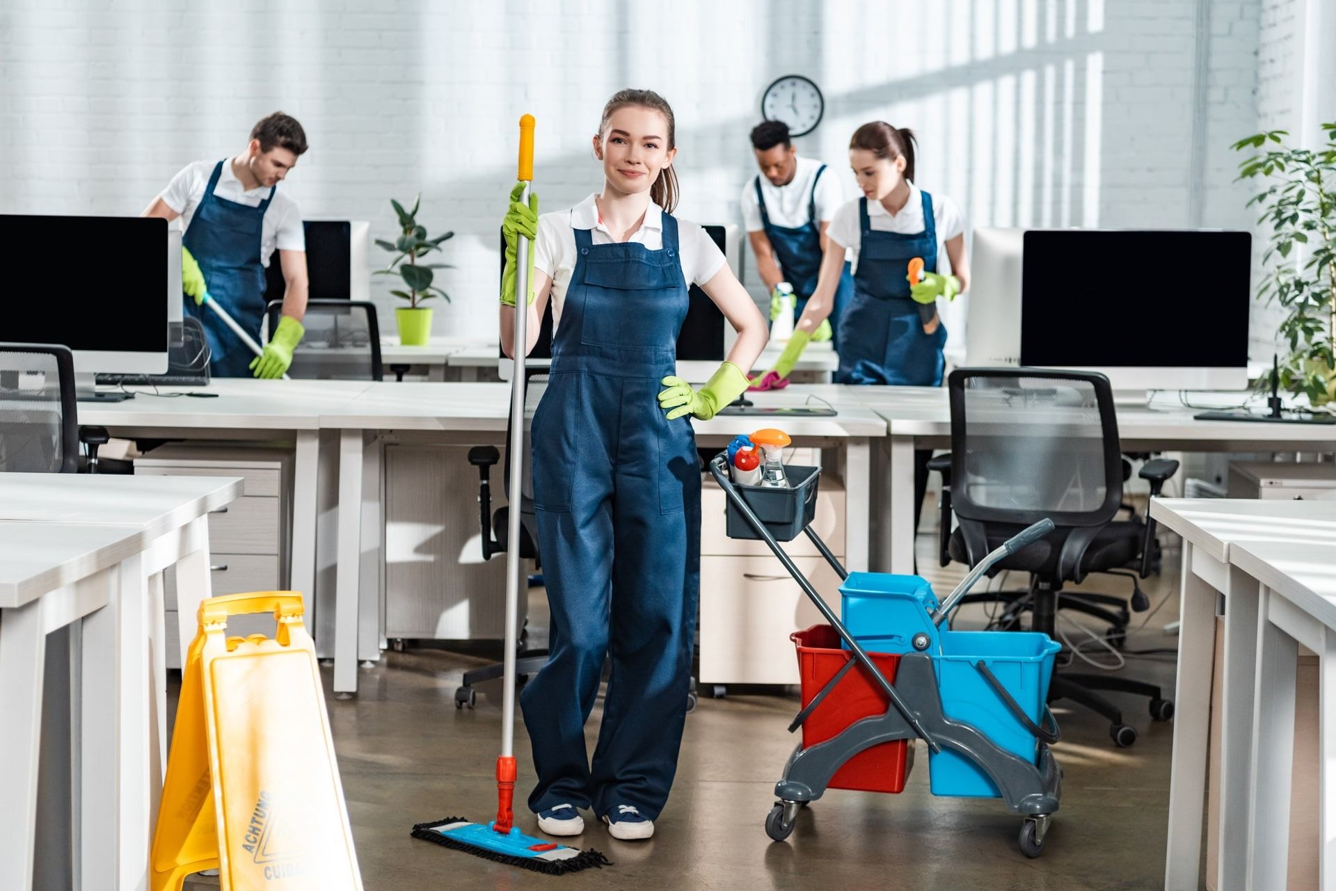 Office cleaning crew working, smiling woman with mop in foreground, desks and equipment in background.