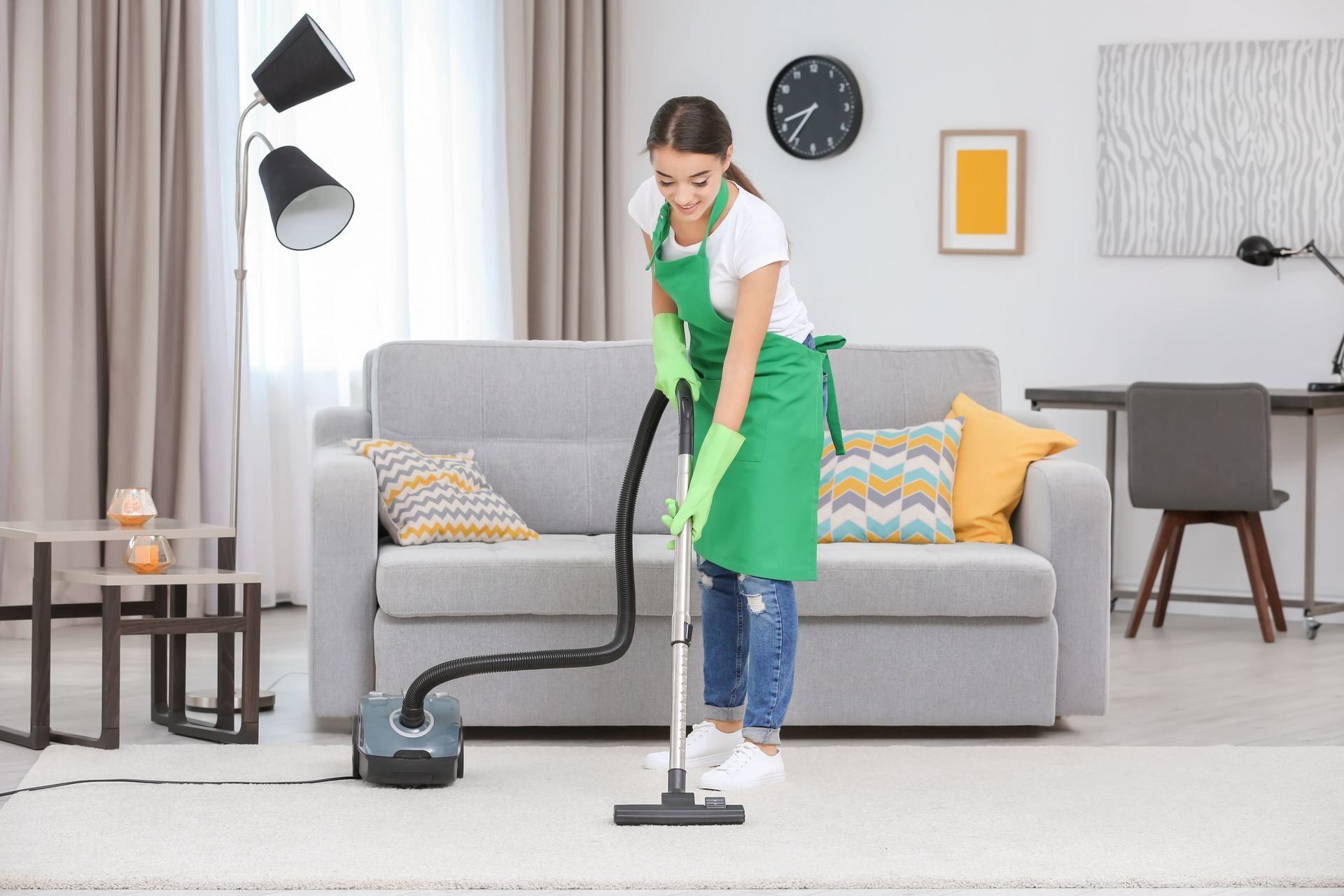 Woman vacuuming a carpet in a living room. She wears a green apron and gloves.