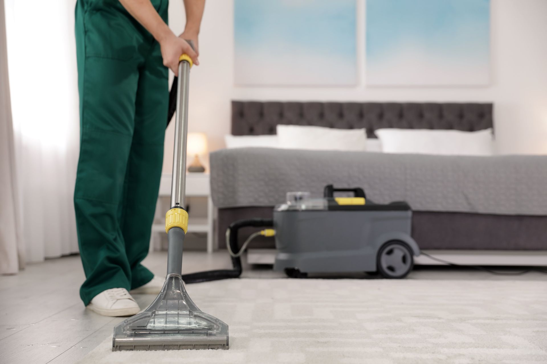 Person using a carpet cleaner in a bedroom, wearing green overalls.