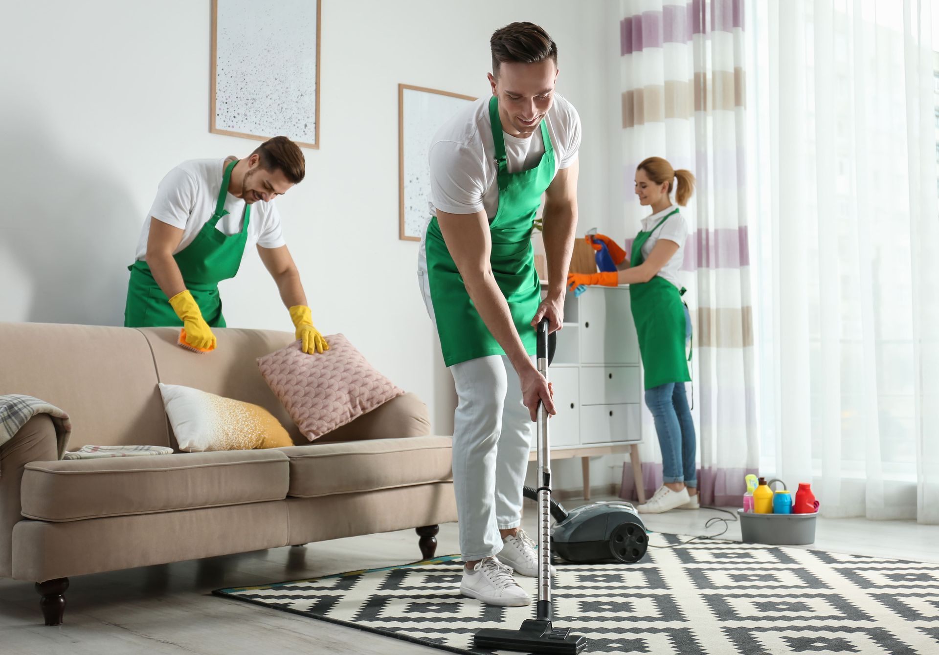 Three people cleaning a living room; one vacuums, two dust and wipe with cleaning supplies.