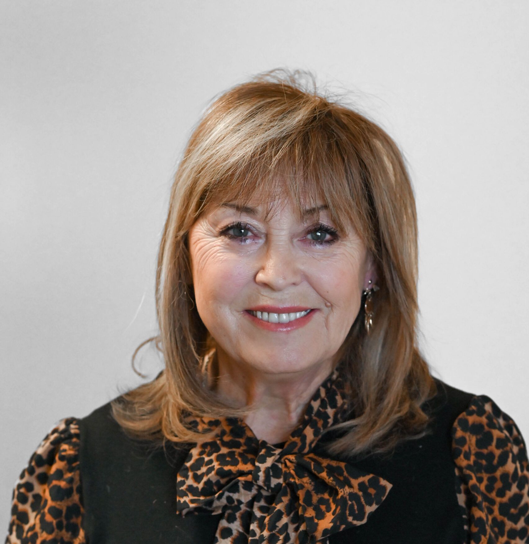 Woman with shoulder-length brown hair, smiling, wearing a patterned blouse and black vest, set against a plain background.