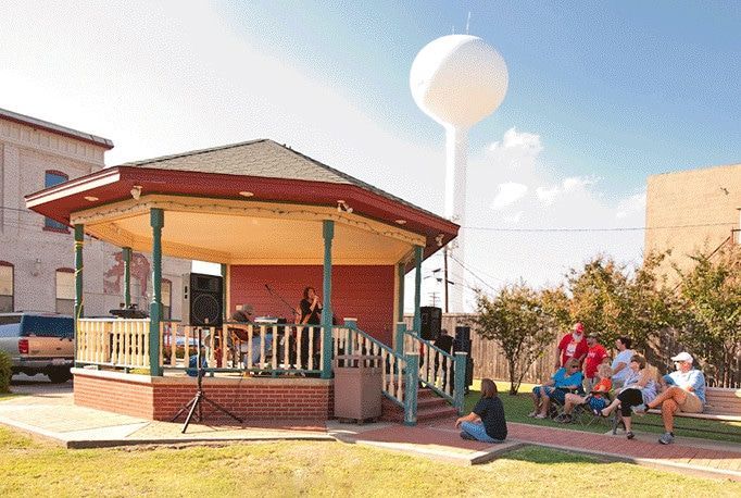 Band performs on a red gazebo stage; a small audience sits on the grass. A water tower is in the background.