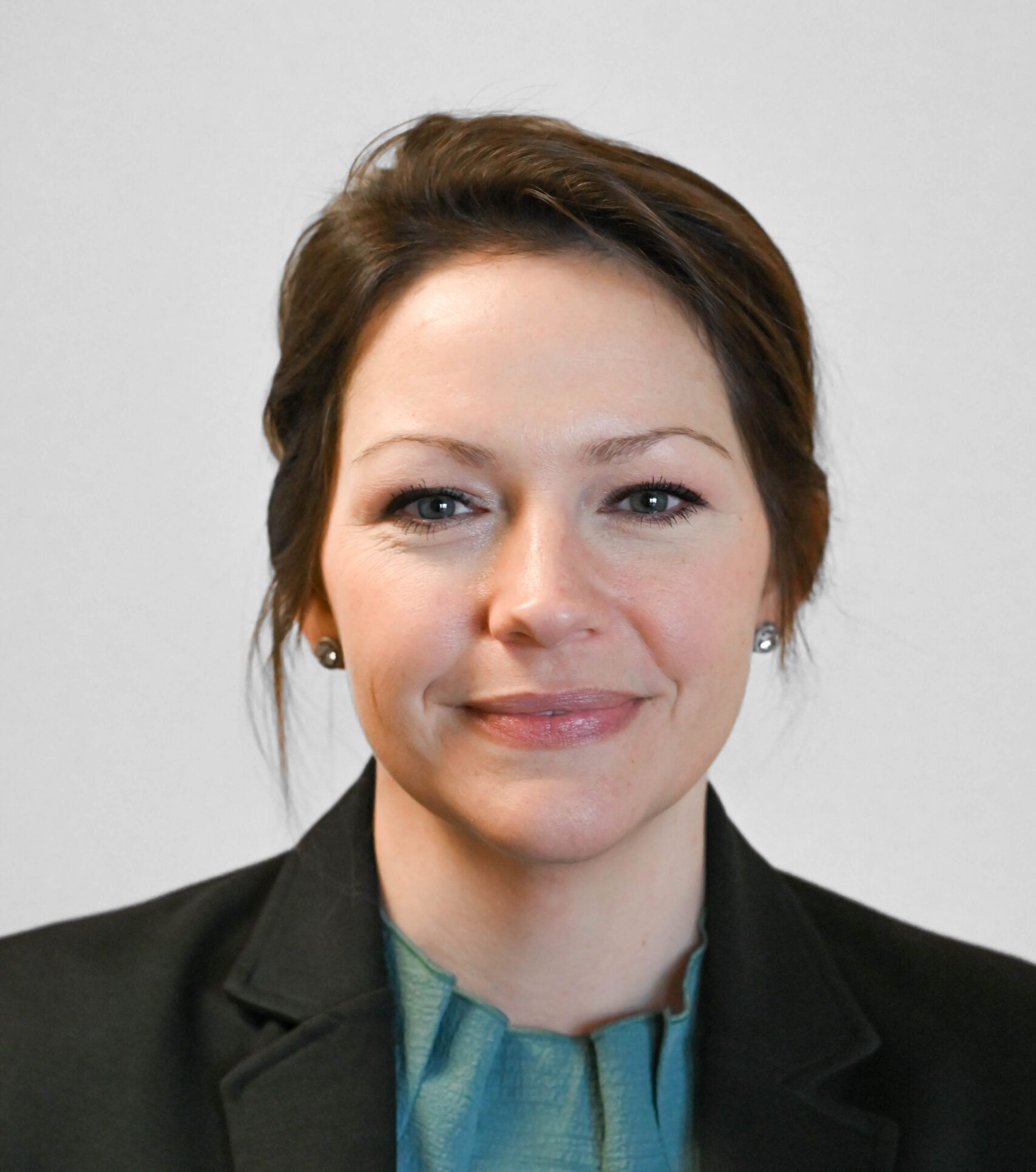 A woman with brown hair wearing a black blazer and teal top smiles at the camera against a light gray background.