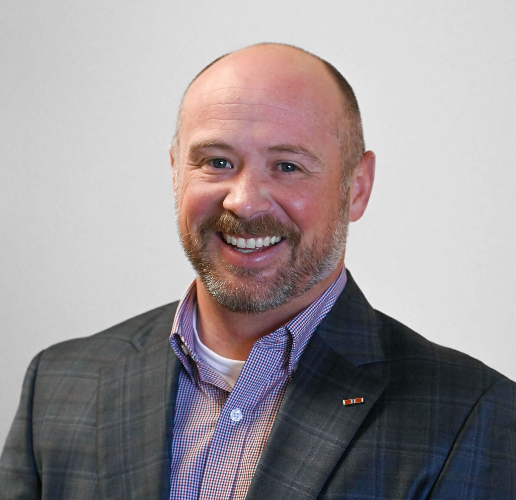Man with a beard smiling, wearing a blazer over a patterned shirt, set against a light gray background.