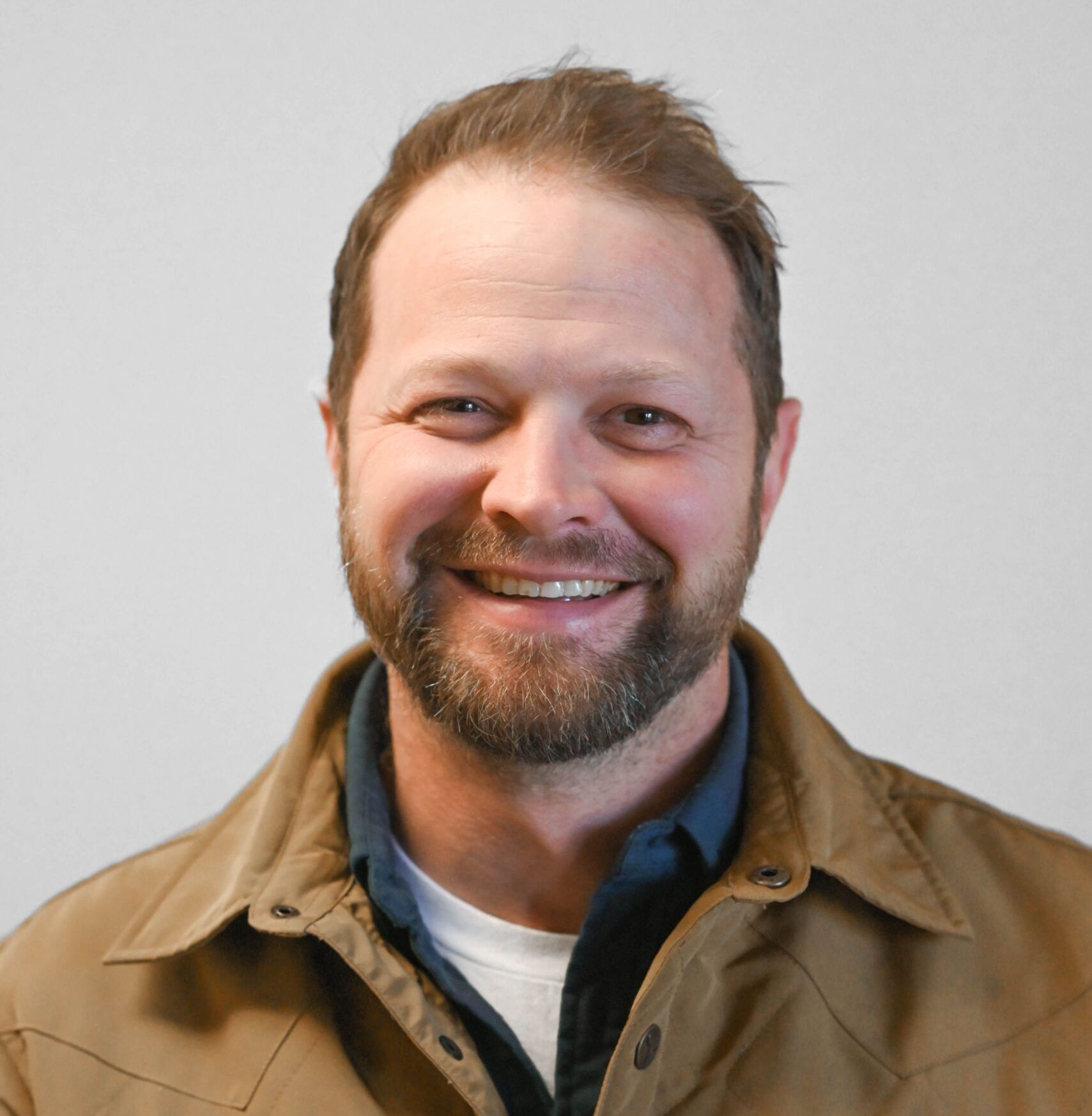 Smiling man with a beard, wearing a brown jacket and blue shirt. He is facing the camera in front of a white background.