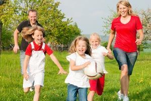 Family of five running and laughing in a grassy field on a sunny day. The children are holding a soccer ball.