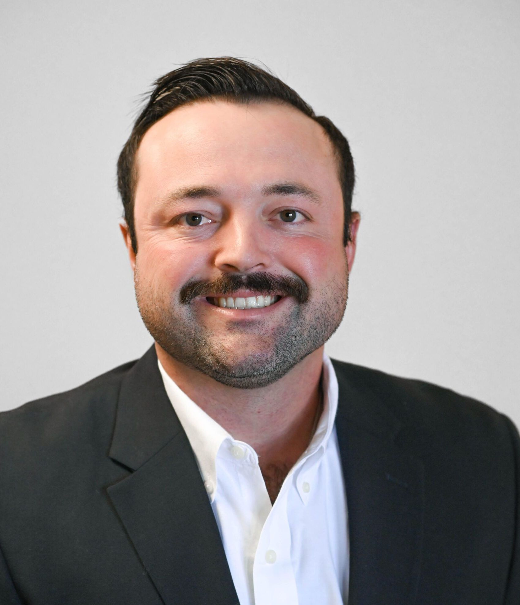 Man in a suit smiling, with a mustache and short dark hair, against a light gray background.