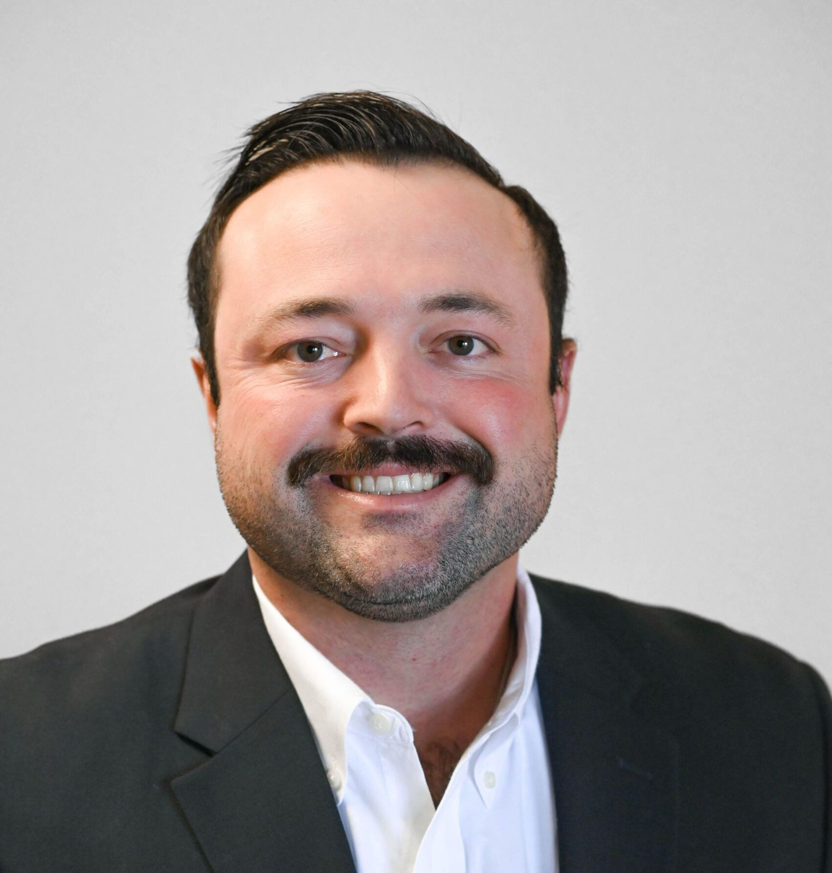 Man in a suit smiling, with a mustache and short dark hair, against a light gray background.