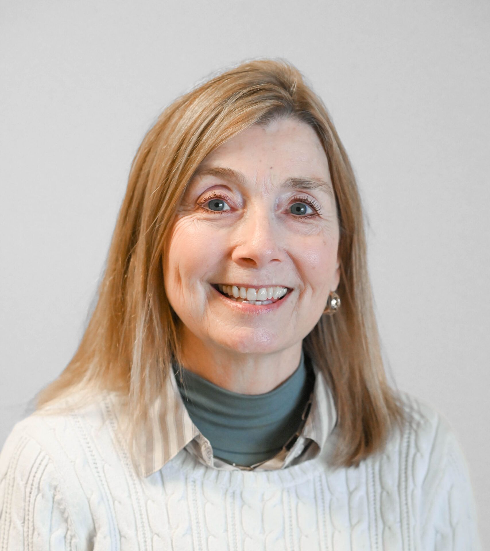 Smiling woman with shoulder-length blonde hair, wearing a light sweater over a teal shirt, against a plain background.