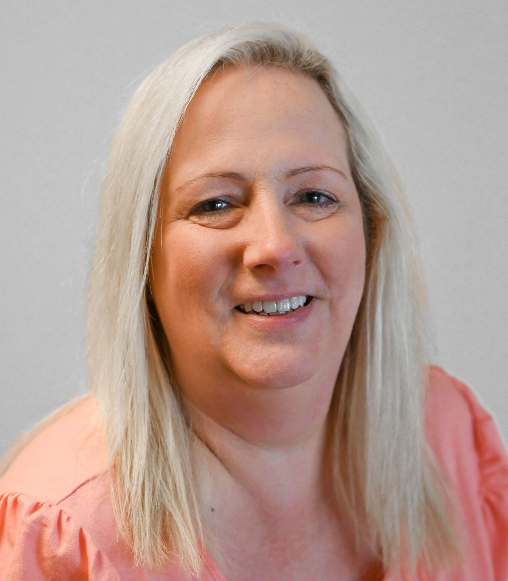 A middle-aged woman with blonde hair and a pink shirt smiles at the camera against a gray background.