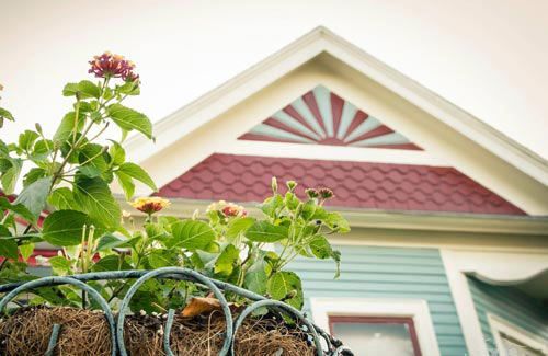 Close-up of a teal house with red roof trim and a flower basket in the foreground. The sunburst design above the windows is visible.