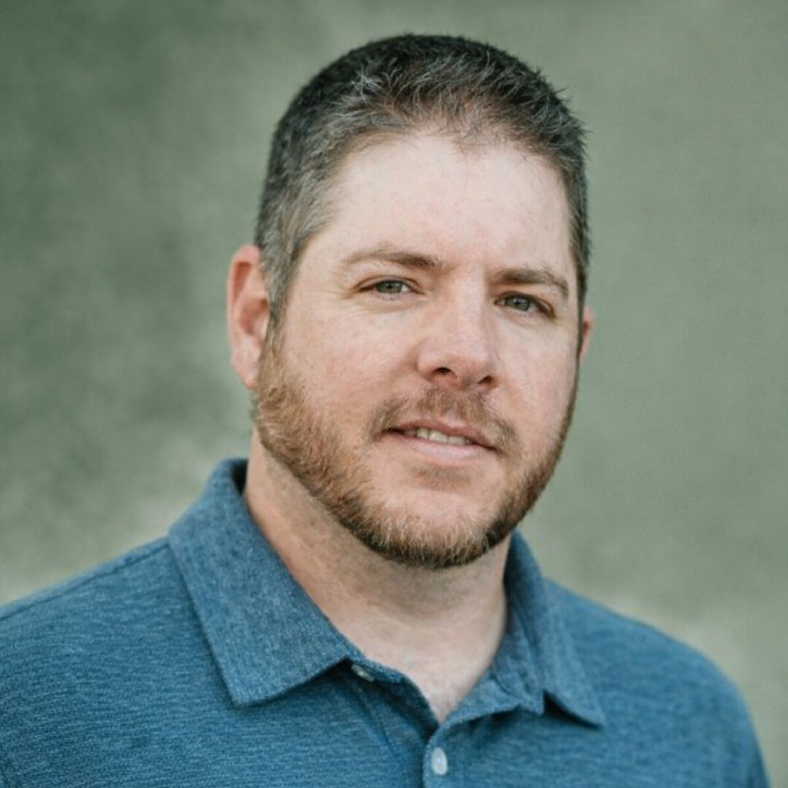 Man with a beard smiling, wearing a blazer over a patterned shirt, set against a light gray background.