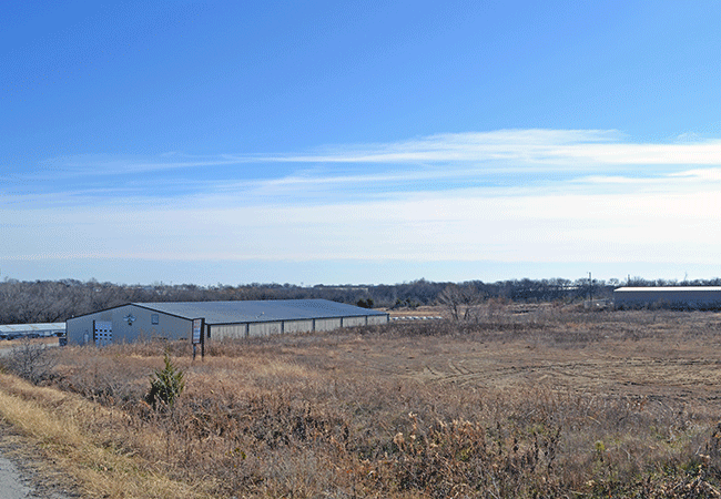 A large metal building sits in a field of dry grass under a clear blue sky. A line of trees is visible in the distance.