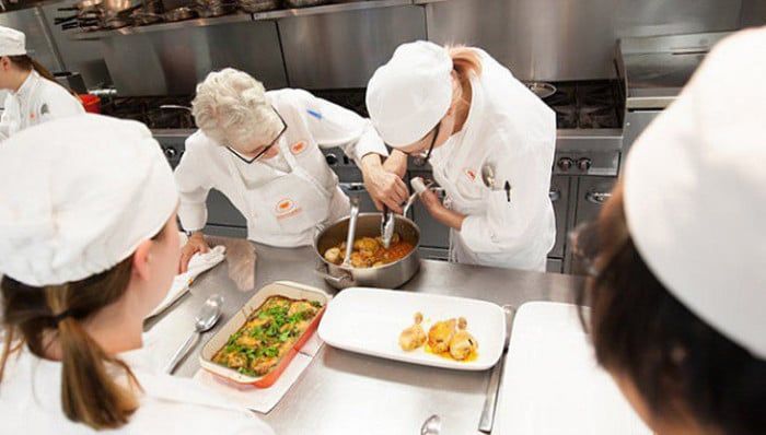 Chefs in white uniforms working in a commercial kitchen, stirring food in a pot. Other dishes are plated on the counter.