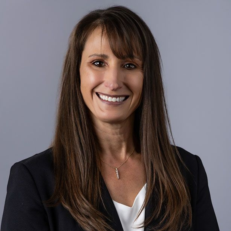 Smiling woman with shoulder-length blonde hair, wearing a light sweater over a teal shirt, against a plain background.