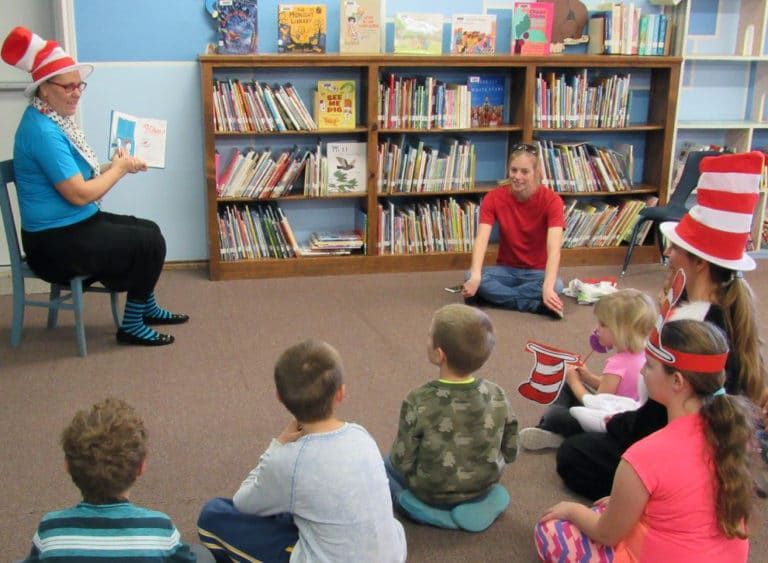 A librarian dressed as the Cat in the Hat reads to children in a library. Some children wear Cat in the Hat hats, everyone is sitting on the floor.