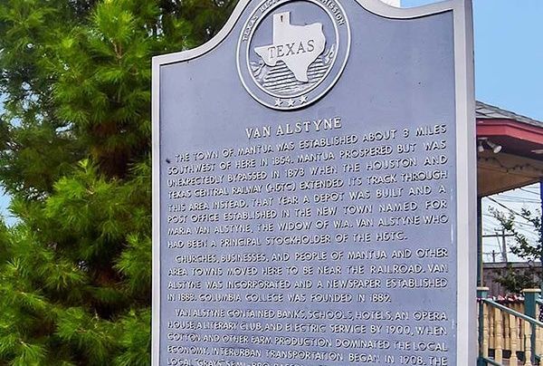 Historical marker for Van Alstyne, Texas, with the state seal at the top. It's outdoors, next to a green bush.