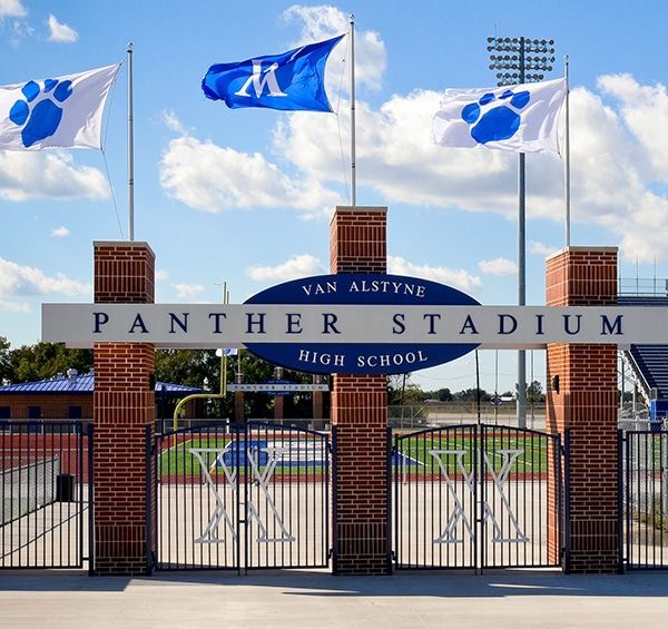 Entrance to Van Alstyne Panther Stadium. Brick columns and black gates frame the stadium's name. Blue and white flags fly above.