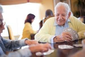 Two elderly men playing cards at a table. One smiles, holding cards. Others sit in the background.