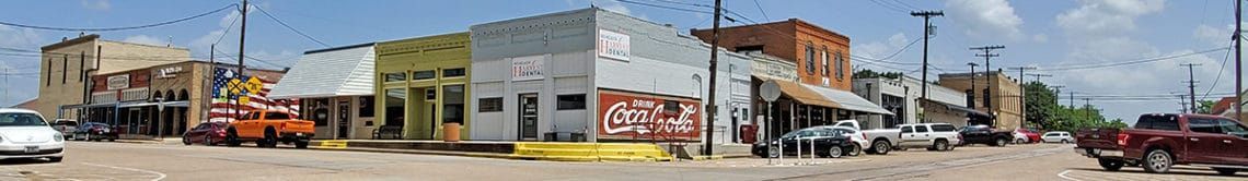 A street scene with historic buildings and cars on a sunny day. The buildings are mostly one-story.