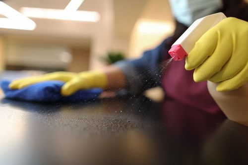 A person wearing a mask and gloves is cleaning a counter with a cloth and spray bottle