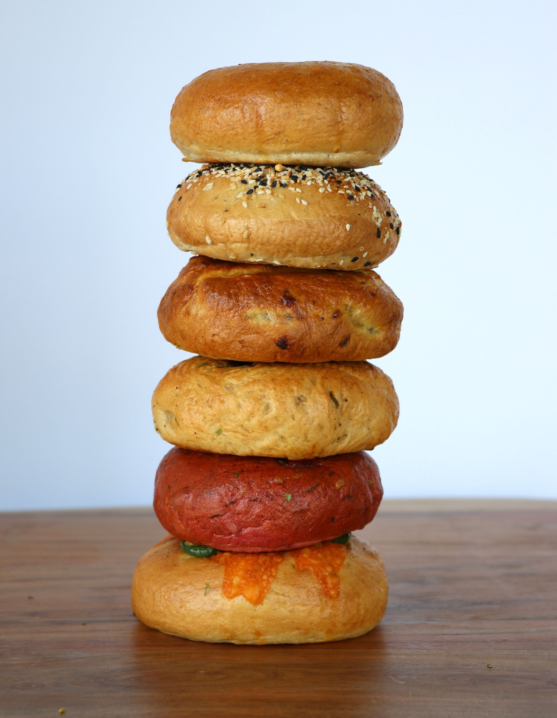 A stack of six bagels of various types and colors piled vertically on a wooden surface against a light background from The Forge Bagel