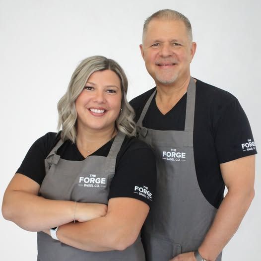 Glen and Rachelle wearing matching aprons, smiling at the camera. They stand in front of a white background.