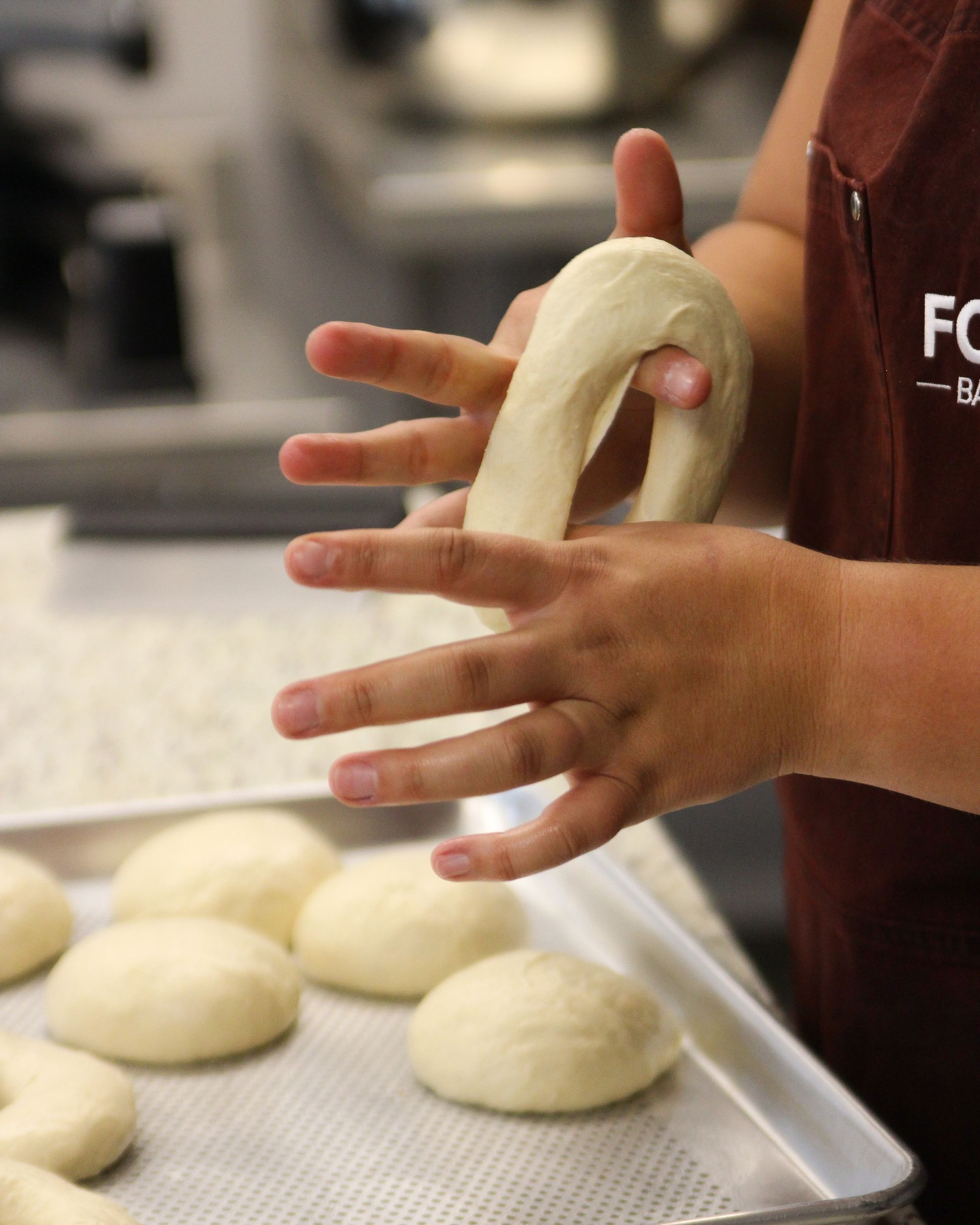 Hands shaping dough into a Polishire Bagel, with a tray of dough balls in the foreground.
