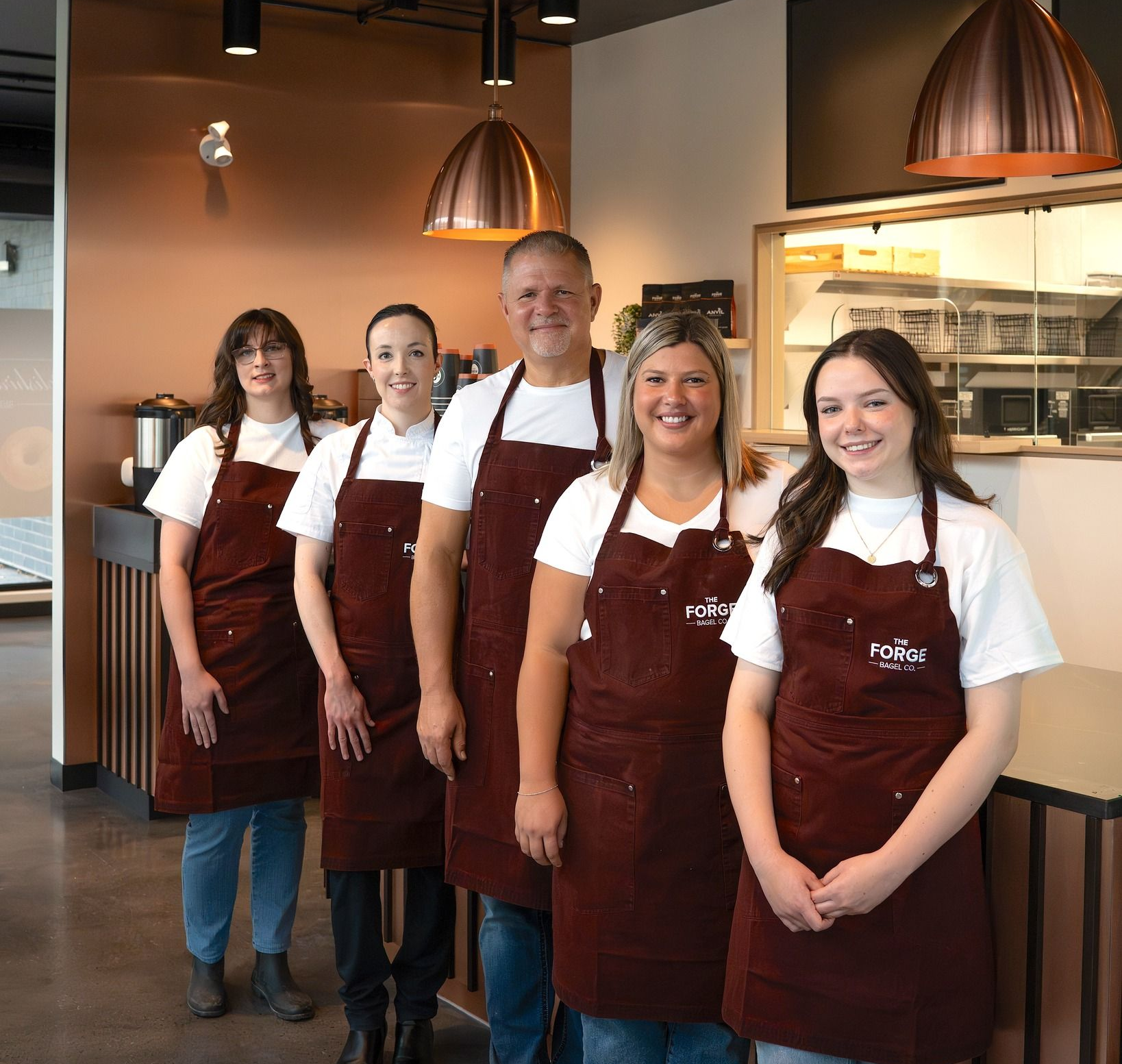 The Forge Bagel Team in brown aprons and white shirts stand in a cafe setting.