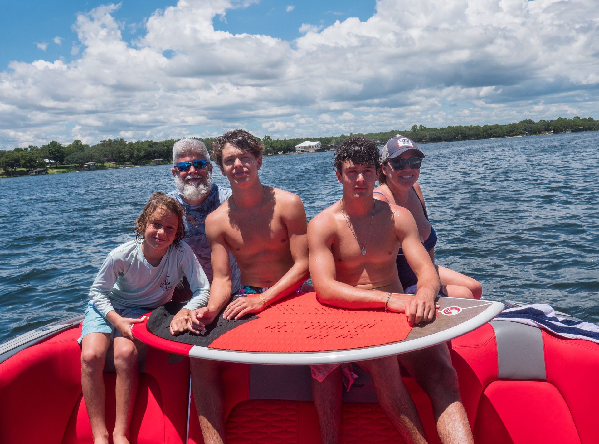 A family is sitting on a boat holding a surfboard.